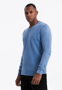 Suéter de punto azul claro con escote en V y puños y dobladillo acanalados, que presenta una textura suave y un diseño ajustado. Llevado sobre pantalones negros.