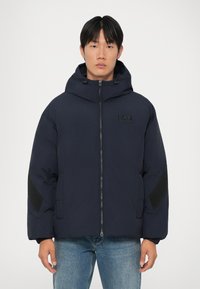 Casaco puffer azul marinho com capuz, fecho de correr na frente e bolsos laterais. Apresenta realces texturizados pretos nos braços e um emblema no peito.
