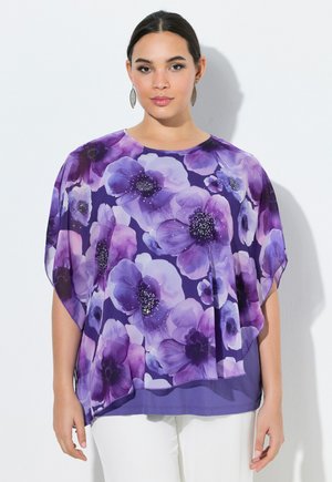 Kvinde iført en løs lilla bluse med store blomsterprint i nuancer af lilla og hvid, kombineret med hvide bukser, stående mod en hvid baggrund.
