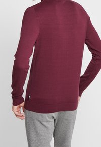 Maglione a maglia marrone con maniche lunghe e orlo a coste. Texture a maglia piatta, design aderente e una piccola etichetta del marchio in basso a sinistra.