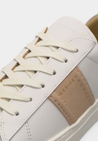Baskets blanches avec panneau latéral en cuir beige, lacets couleur crème et logo "NUBIKK" doré sur le côté.