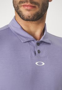 Polo shirt violet avec un col, trois boutons et un logo brodé. Texture de tissu douce avec une finition lisse.