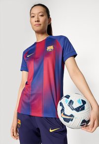 FC Barcelonas tröja i röd och blå ränder, kortärmad, med en märke och Nike-logotyp. Spelaren håller i en vit, svart och blå fotboll.