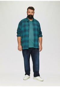 Turquoise t-shirt met korte mouwen, gedragen onder een geruit overhemd in donker teal en zwart, gecombineerd met donkere jeans en witte sneakers.