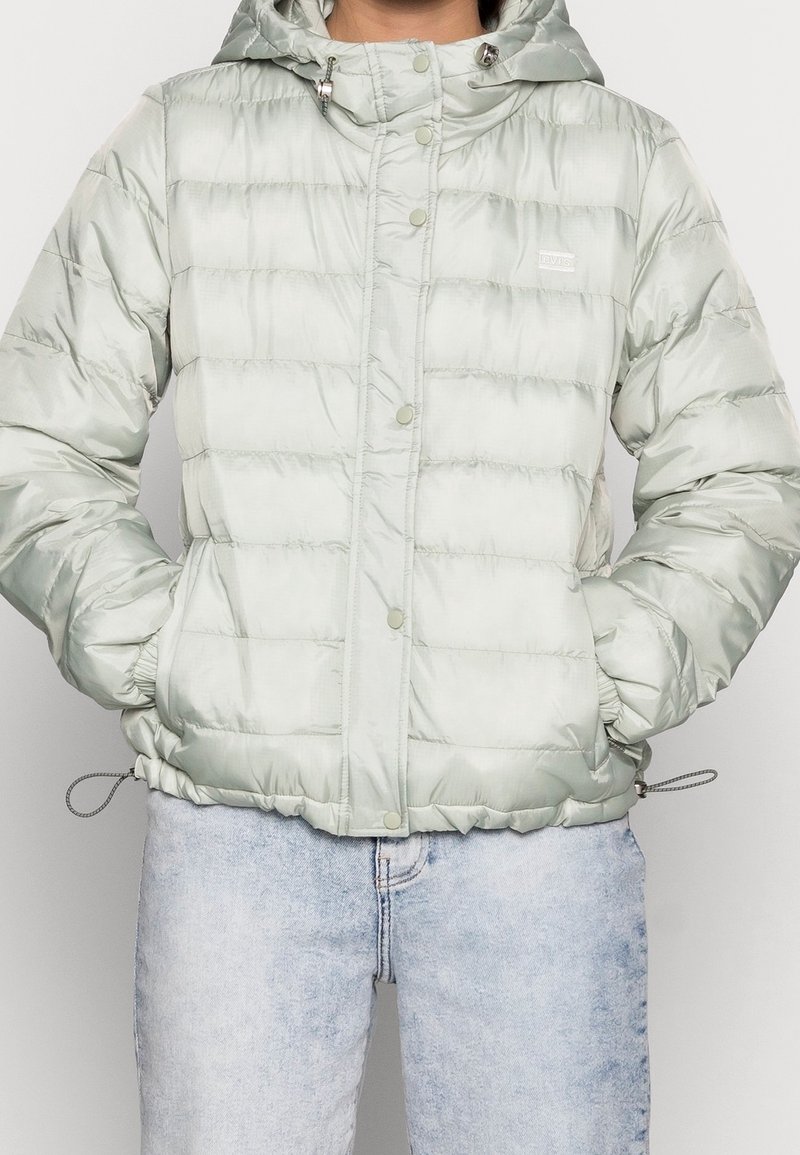 Chaqueta puffy verde claro con capucha, diseñada con un cierre frontal a presión, puños elásticos y dobladillo ajustable con cordón. Textura suave y brillante.