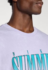 Lyse lilla t-shirt med rund halsudskæring, der har stor turkis "SUMMER" tekst. Blødt bomuldsstof med en subtil tekstur. Afslappet design.