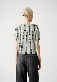 Top de manga corta verde claro con diseño de hojas en negro, con un corte holgado y detalle de espalda fruncida, combinado con unos jeans gris oscuro.