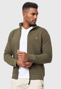 Chaqueta verde oliva con cremallera y un patrón texturizado, puños acanalados y un parche de logo redondo en el pecho. Llevada sobre una camiseta blanca.