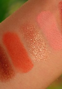 Vier Farbproben von Make-up auf der Haut: dunkelbraun mit Schimmer, mattes Orange, schimmerndes Gold und zwei matte Rosatöne, variierend in Deckkraft und Textur.