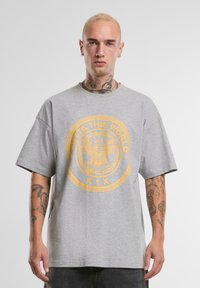 Grå oversized t-skjorte laget av bomull, med et gullsirkulært grafisk motiv med teksten "OUTTA THIS WORLD" og en ørnemblem. Korte ermer.