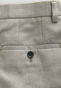 Light beige, textured fabric with a button closure on a back pocket. Features stitched detailing and a clean waistband design.