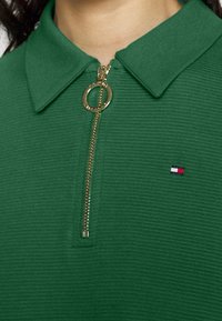 Zelená polo tričko na zip vyrobené z texturované látky, s límečkem a kulatým zlatým táhlem zipu. Malé logo v červené, bílé a modré barvě.