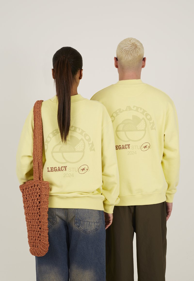 Lyse gule hettegensere med en stor brun grafikk på baksiden. En modell bærer en brun, heklet veske. Buksene er løstsittende og mørke.