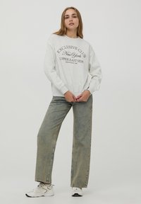 Felpa grigia chiara con la scritta "EXCLUSIVE CLUB New York UPPER EAST SIDE" stampata in un colore scuro. Abbinata a jeans larghi verde sbiadito e sneakers.