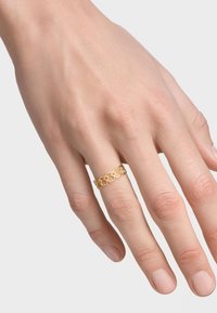 Złoty pierścionek o splocie, założony na nagą dłoń. Projekt wyróżnia się ażurową konstrukcją oraz skomplikowanym, powtarzającym się kształtem.