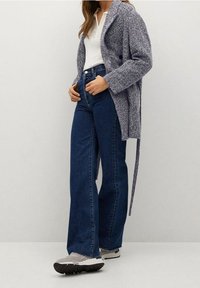 Cardigan grigio a maglia con apertura frontale, abbinato a jeans blu a vita alta e gamba larga e sneaker grigi. Vestibilità strutturata e comoda con maniche lunghe.