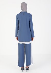 Tunica lunga blu con orlo bianco, abbinata a pantaloni larghi coordinati. Presenta una scollatura a chiave sul retro ed è completata da un hijab grigio chiaro.