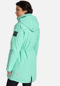 Mėtų žalios spalvos, vandeniui atspari striukė su raišteliu liemens srityje, gobtuvu ir atspindinčiais akcentais; kairiajame rankovėje yra juodas užrašas.