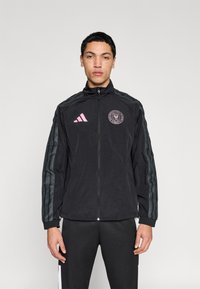 Μαύρο αθλητικό μπουφάν με φερμουάρ, με ροζ λογότυπο και ριγέ γκρι διακοσμητικά στις μανσέτες. Το υλικό φαίνεται ελαφρύ και λείο.