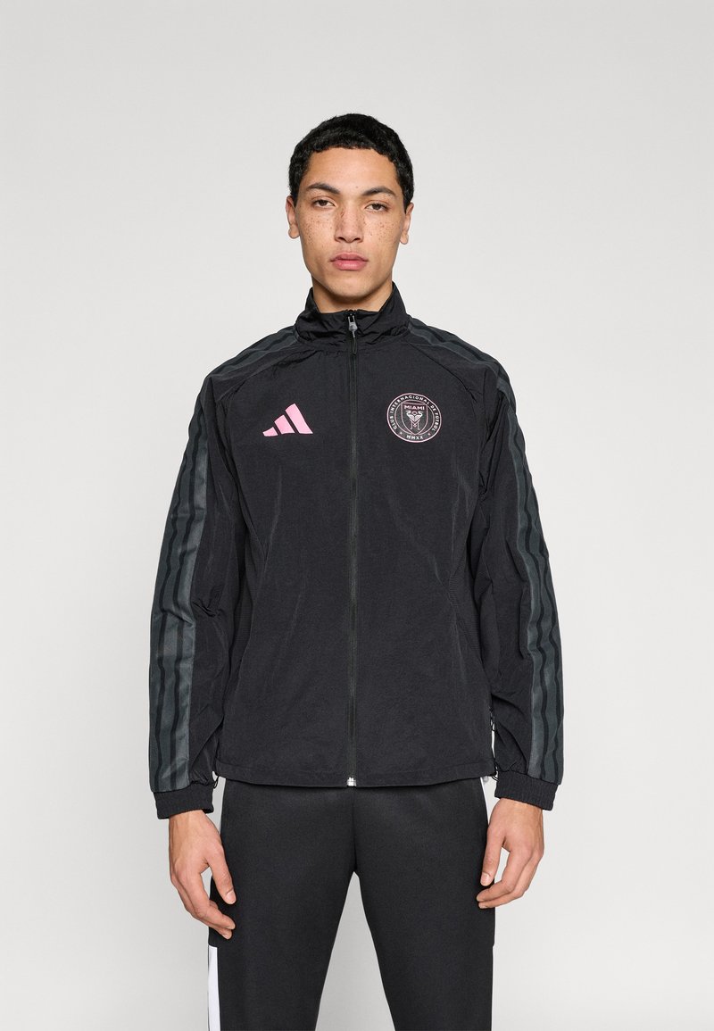 Μαύρο αθλητικό μπουφάν με φερμουάρ, με ροζ λογότυπο και ριγέ γκρι διακοσμητικά στις μανσέτες. Το υλικό φαίνεται ελαφρύ και λείο.