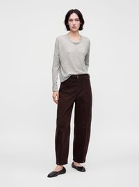 Jeune personne debout portant une chemise à manches longues gris clair, un pantalon ample marron foncé et des chaussures plates noires sur un fond clair uni.