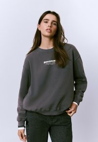 Sweatshirt gris avec un col rond, arborant un texte de logo blanc. Il a une coupe ample et est associé à un jean en denim foncé.