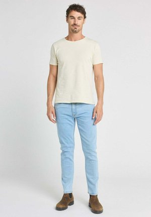 Homme debout portant un t-shirt beige uni, un jean bleu clair et des bottines marron, sur un fond blanc uni.