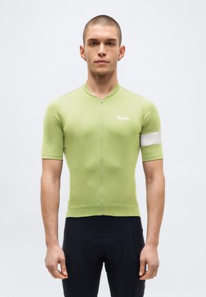 Homme portant un maillot de cyclisme vert clair à manches courtes avec un brassard blanc et un cuissard rembourré noir, debout devant un fond uni.
