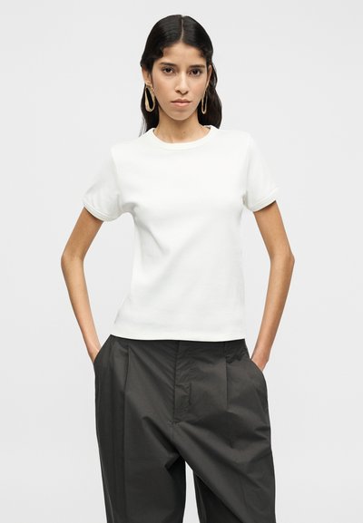 Λευκό κοντομάνικο μπλουζάκι με στρογγυλή λαιμόκοψη, λεία υφή και κλασική εφαρμογή, συνδυασμένο με ψηλόμεσα, σκουρόγρι μαύρα παντελόνια.