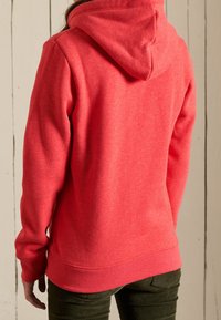 Persona vestida con una sudadera con capucha roja y pantalones oscuros de espaldas a una pared de madera blanca.