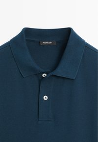 Polo bleu foncé avec un col côtelé et deux boutons blancs sur la patte de boutonnage, l’étiquette indique Fabriqué en Turquie.