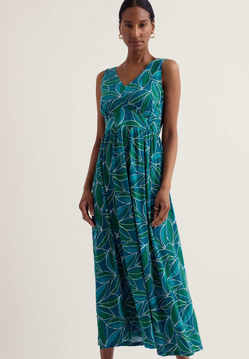 Femme portant une robe maxi verte sans manches avec un motif de feuilles bleues et blanches, debout devant un fond clair uni.