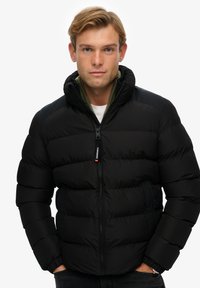 Chaqueta de plumas negra con cierre de cremallera, diseño acolchado, puños elásticos y capucha. El interior cuenta con un forro verde y un detalle del logo en la cremallera.