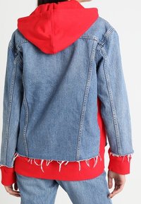 Jeanjacka med röd hoodie-insats, fransad nederkant och tvåfärgad design. Har en avslappnad passform och synliga sömnadsdetaljer.