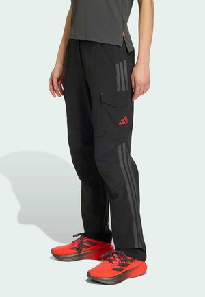 Person iført sorte bukser med sidelommer og grå striber, kombineret med lyserøde og sorte sportssko, stående på en lys baggrund.