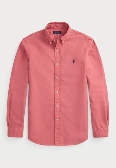 Gaiši rozā krekls ar gariem piedurknēm, apkakli un mazu zilu logo kreisajā krūšu daļā. Kokvilnas audums ar gludu tekstūru.