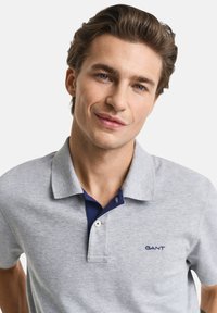 Šedé polo tričko s námořnickým límcem, klasický design, krátké rukávy a modré logo GANT na levé hrudi. Měkká bavlněná struktura.