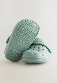 Sabots vert clair en matériau semblable au caoutchouc, dotés d'une sangle, de trous de ventilation et d'une semelle extérieure texturée et à motif pour une meilleure adhérence.