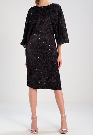 Frau trägt ein schwarzes knielanges Kleid mit weißen sternförmigen Punkten und silbernen Sandalen mit hohem Absatz, steht vor einem einfarbigen Hintergrund.