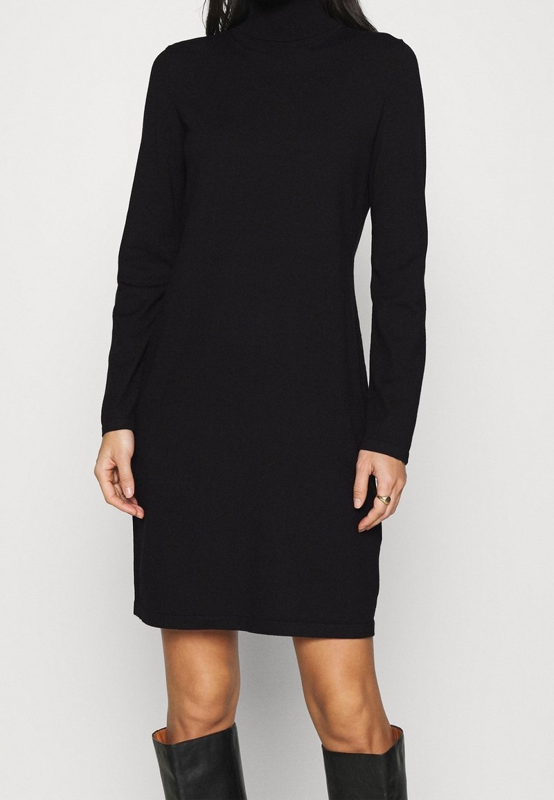 Femme portant une robe noire simple à manches longues et col roulé, associée à des bottes noires montantes, debout devant un fond blanc uni.