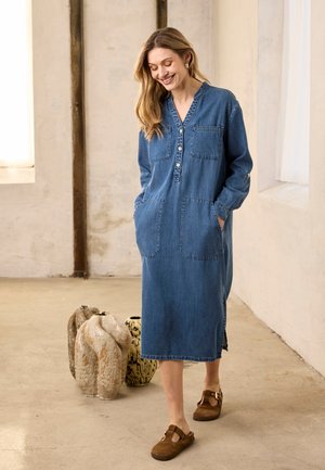 Nő sétál beltéren, kék farmer hosszú ujjú ruhát viselve, foltzsebekkel és barna slip-on cipőben, mosolyogva és lefelé nézve.