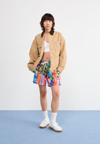 Béžová bunda s golierom na bielom crop top, skombinovaná s živými viacfarebnými šortkami s kvetinovým a prúžkovaným vzorom a bielymi teniskami.