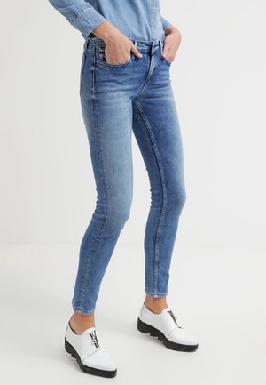 Osoba ubrana w niebieskie obcisłe dżinsy, jasno niebieską koszulę na guziki i białe buty na zamek, stojąca z rękami w kieszeniach.