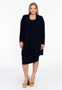 Navyblauer dreiteiliger Anzug bestehend aus einem langen Blazer, einem taillierten Kleid und hochhackigen Plateausandalen mit goldenen Akzenten. Glatte Stoffstruktur.