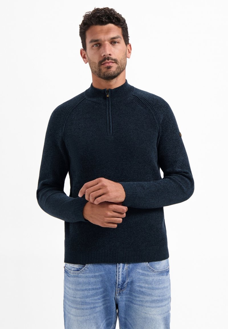 Donkerblauwe gebreide trui met ritssluiting en ribbeltextuur, raglanmouwen en een klein logo op de linkerarm. Draagt over een blauwe spijkerbroek.