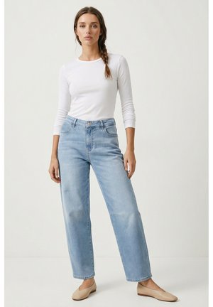 Frau mit langen braunen Haaren in einem Zopf trägt ein weißes Langarmshirt, hellblaue hoch geschnittene Jeans und beige Ballerinas und steht drinnen.