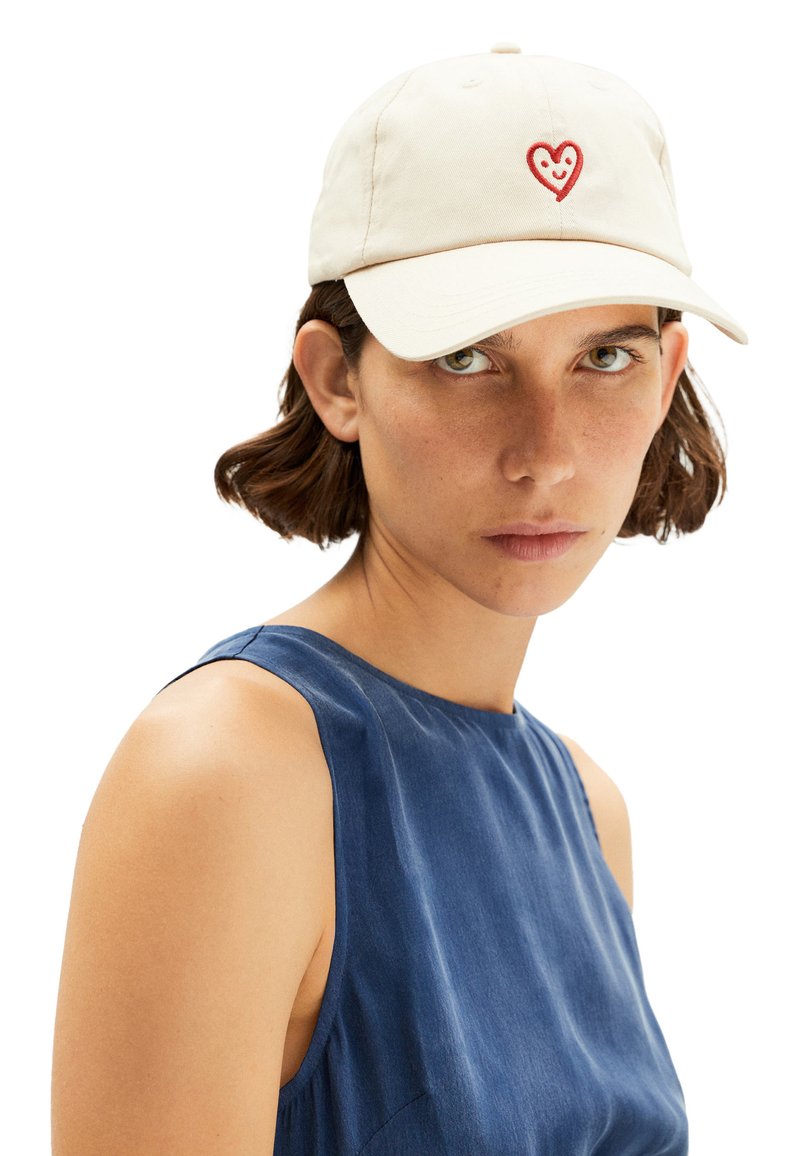 Femme aux cheveux bruns courts portant une casquette de baseball crème avec un motif cœur rouge et un top bleu sans manches regardant devant elle.