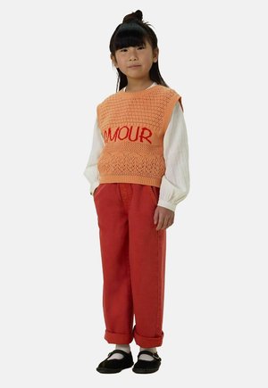 Orange gestrickte Weste mit "AMOUR" in Rot, über einem weißen Langarmshirt getragen. Rote Hose und schwarze Schuhe runden das Outfit ab.