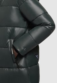 Donkergroen pufferjack met glanzende textuur, gequilt ontwerp en zijzakken. Inclusief merketiketten bij de linker mouw en zak.