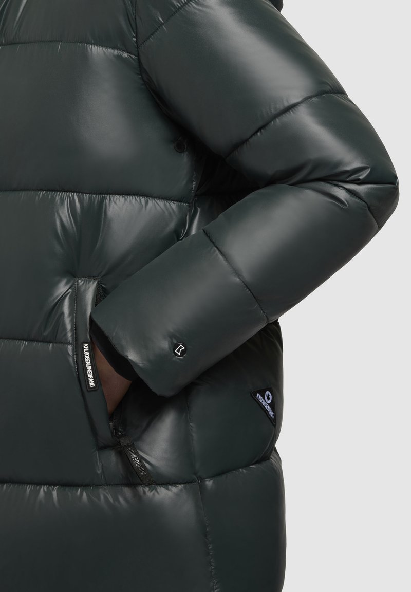 Giacca puffera verde scuro con texture lucida, design trapuntato e tasche laterali. Include etichette di marca vicino al braccio sinistro e alla tasca.